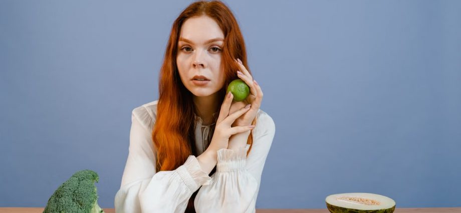 Woman with red hair holding lime surrounded by fresh produce on table.